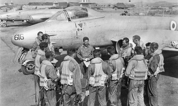  Pilots of No.77 Squadron RAAF, being briefed by their Commanding Officer, Squadron Leader Richard (Dick) Cresswell (centre, facing camera), on the Kimpo airfield tarmac in front of Meteor aircraft A77-616, prior to a mission over North Korea in 1951. Flight Lieutenant Maxwell (Max) Scannell, RAF is second from the left in the semicircle. Australian War Memorial JK0024 