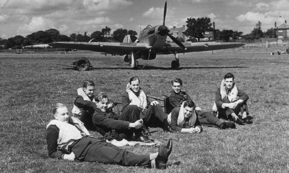 Group of pilots from 'B' Flight, Number 32 Squadron, resting on the grass in front of one of their Hurricanes at Royal Air Force Station Hawkinge.  This is during the Battle of Britain. L-R: Pilot Officer RF Smythe, Pilot Officer K Gillman, Pilot Officer JE Proctor, Flight Lieutenant PM Brothers, Pilot Officer DH Grice, Pilot Officer PM Gardner, Pilot Officer AF Eckford. Image credit: Air Force Museum of New Zealand. 
