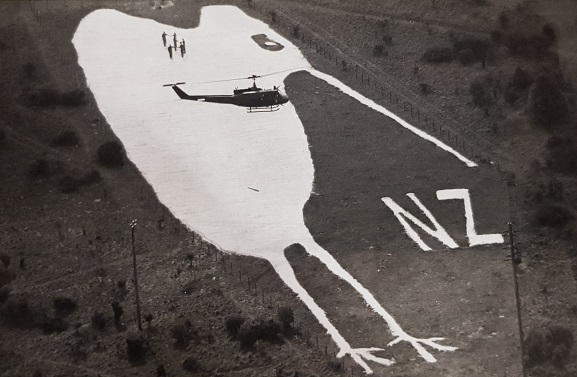 Members of the 3 Squadron Helimeet 90 team standing on the famous chalk Kiwi at Bulford, UK, carved by New Zealand soldiers during World War One. Iroquois NZ3801 hovering overhead. Image: 3 Squadron RNZAF Unit History.
